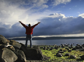 Photo of a man holding his arms up in front of a lake.